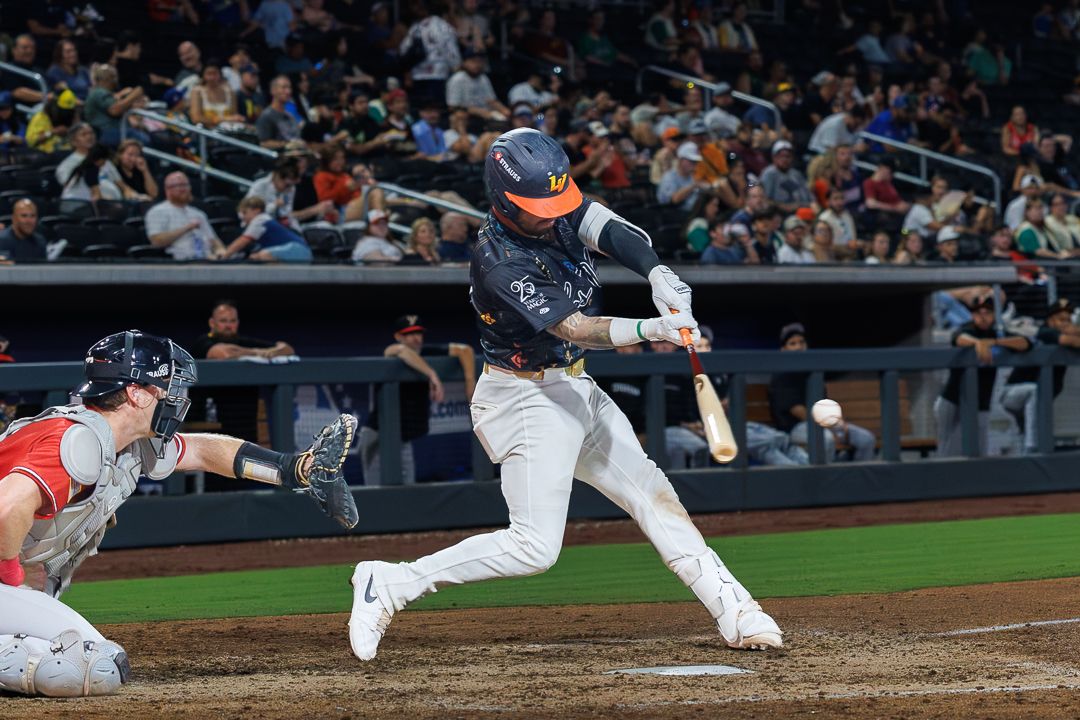 Baseball batter in dark uniform swings at pitch during night game with catcher and crowd in background.