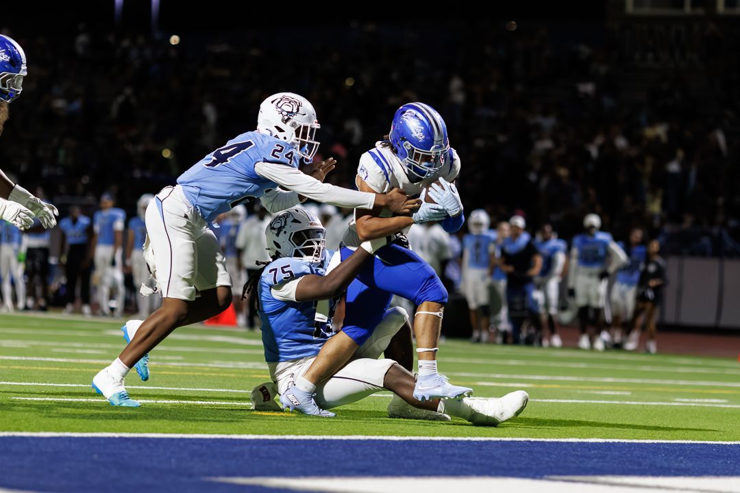 Football player in blue uniform scores touchdown while being tackled by defenders during a night game.