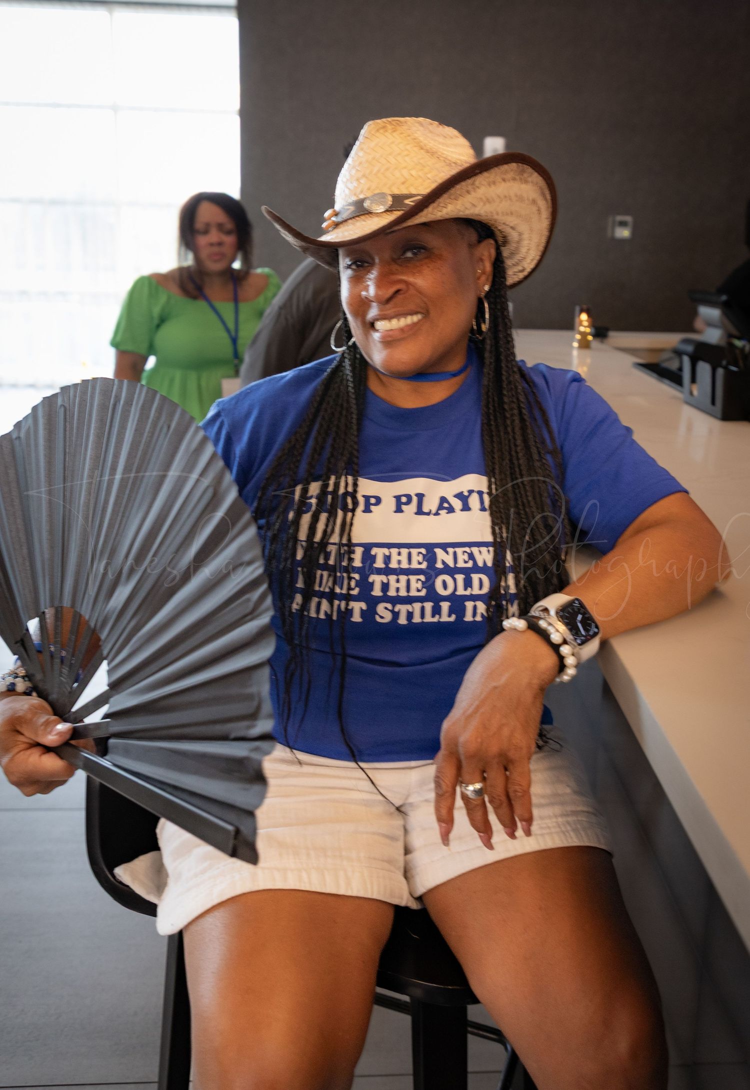Event participant wearing a cowboy hat and blue t-shirt holds a decorative fan while seated indoors.