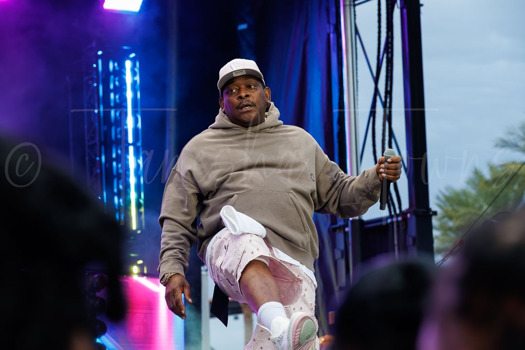 A performer in a beige hoodie and white pants energetically performs on a colorful outdoor festival stage.
