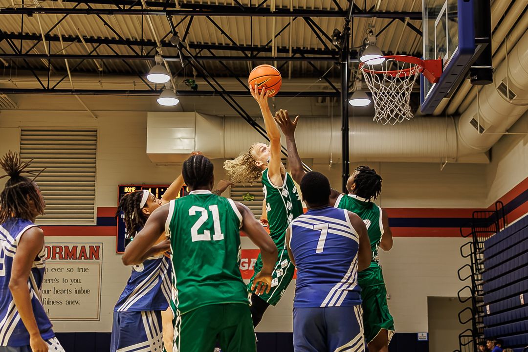Basketball players compete for a rebound near the hoop during an indoor game, with green and blue team uniforms.