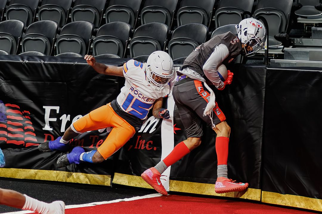 Two football players in orange and blue uniforms battle near the sideline during an indoor arena football game.