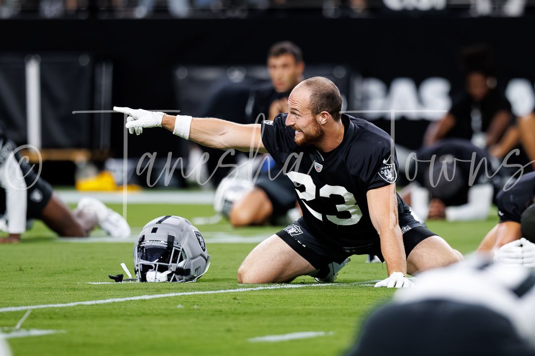 NFL player #23 in black Raiders uniform kneels on field pointing during warmups at Allegiant Stadium.