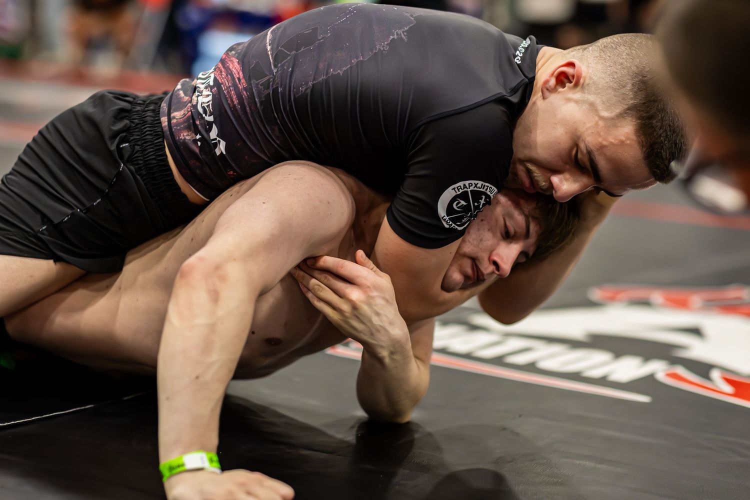 Two athletes competing in a grappling match on the mat during an MMA or BJJ tournament.