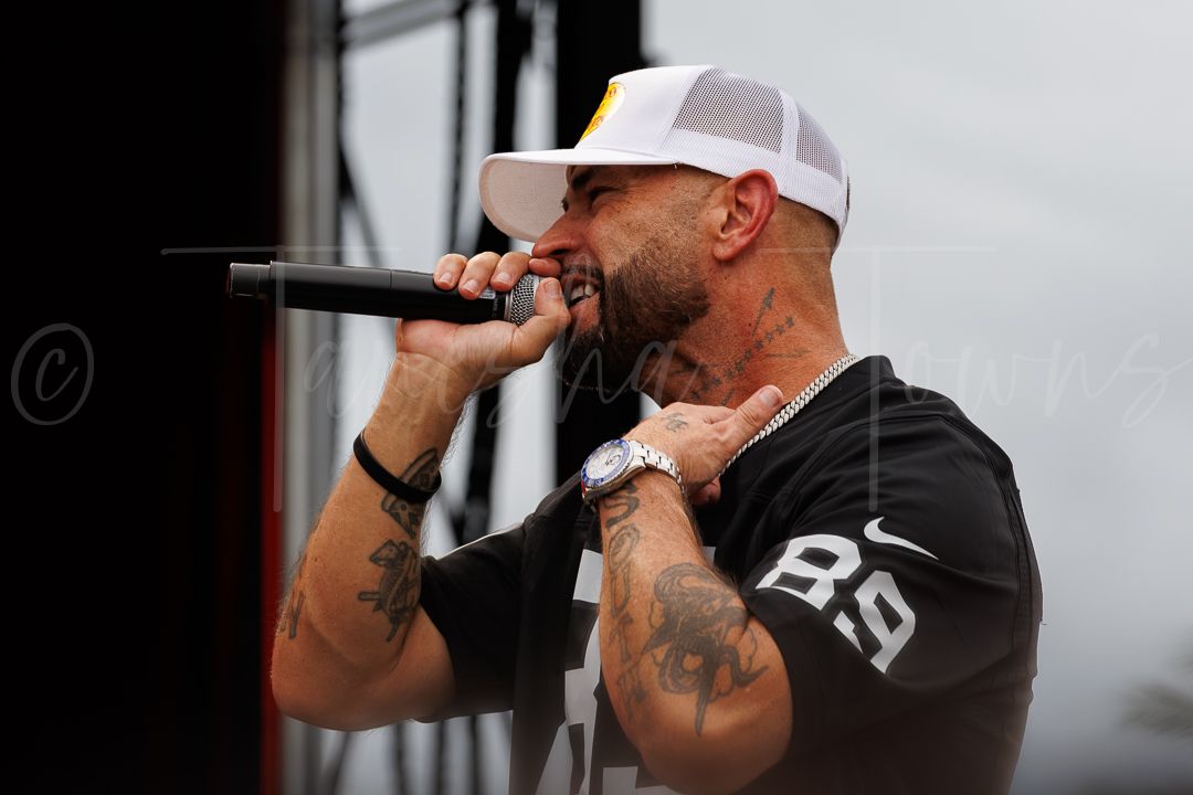 Tattooed male rapper performing on outdoor stage, wearing white snapback cap and black jersey, singing into microphone.