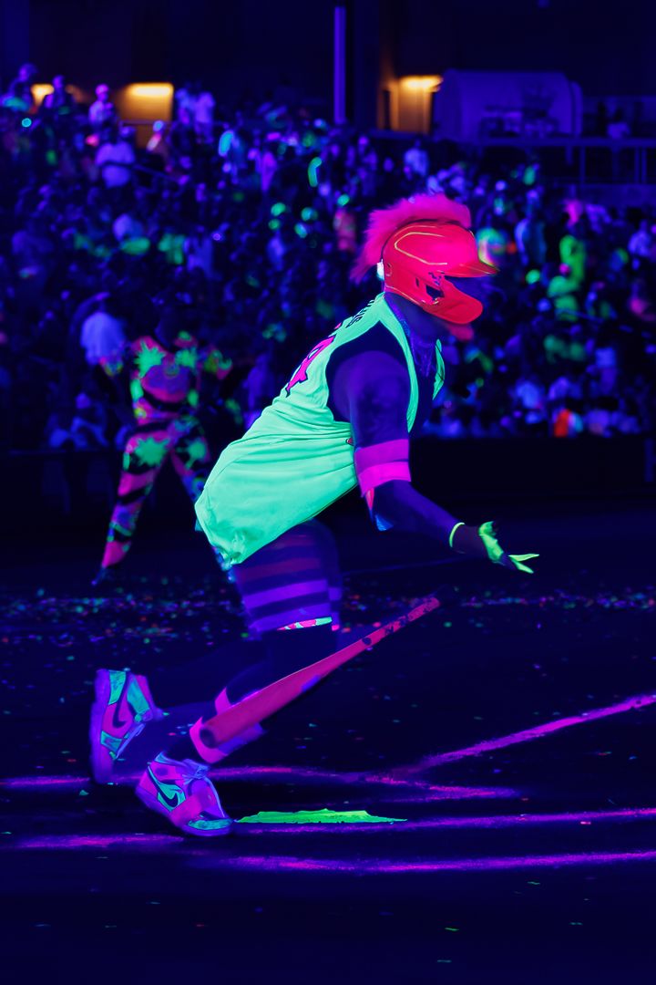 Person in neon UV-reactive outfit with pink helmet performs energetic dance move at blacklight event.