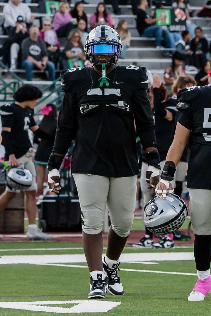 Football player in black uniform and blue visor standing on field during game.