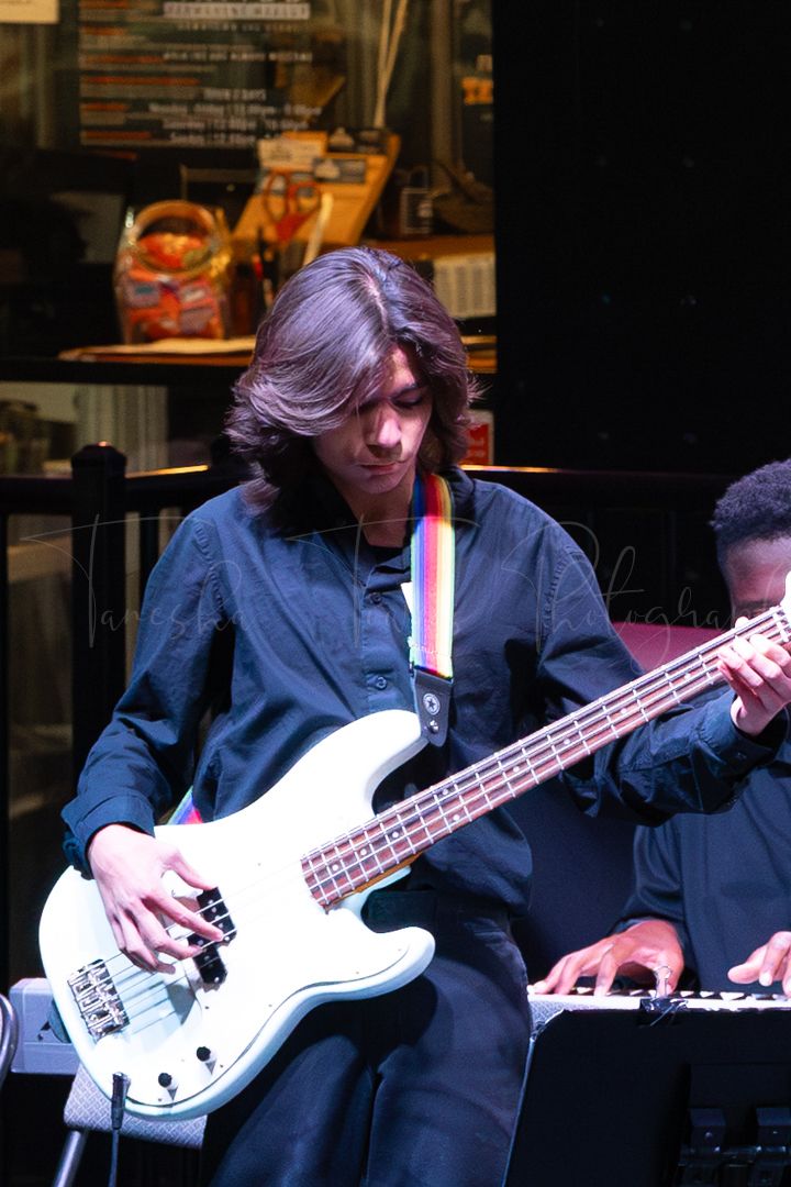 Musician playing a white bass guitar on stage wearing a dark blue shirt.