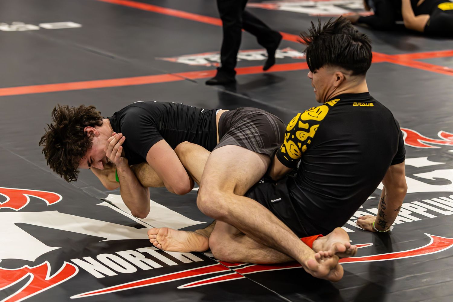 Two athletes compete in a grappling match on a black mat at an MMA or BJJ tournament.