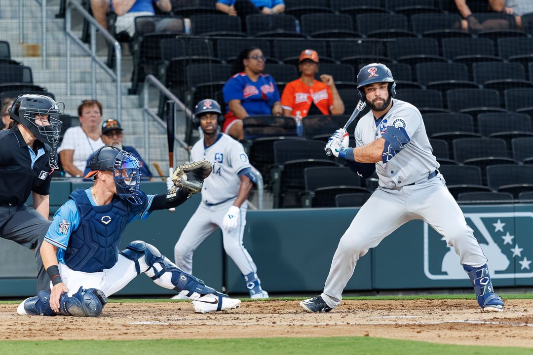 Baseball batter prepares to swing at home plate while catcher and baserunner watch during a minor league game.
