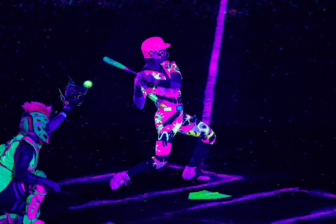 Players in neon glowing outfits play baseball under black lights in a vibrant UV-lit arena.
