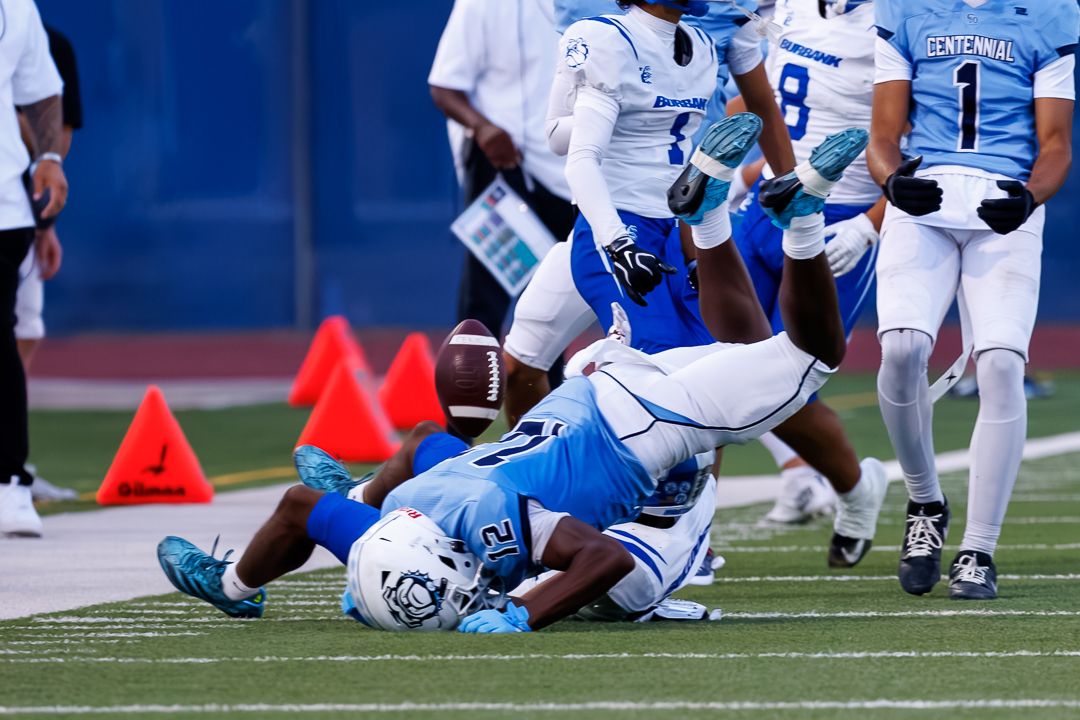 Football players collide on the field during a daytime game with orange cones visible in background.