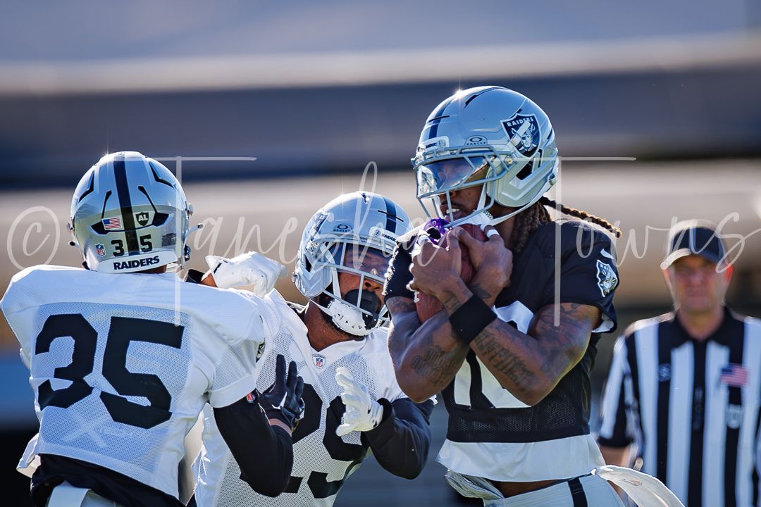 Raiders players in black and white uniforms compete for a football catch during an outdoor practice session with a referee nearby.