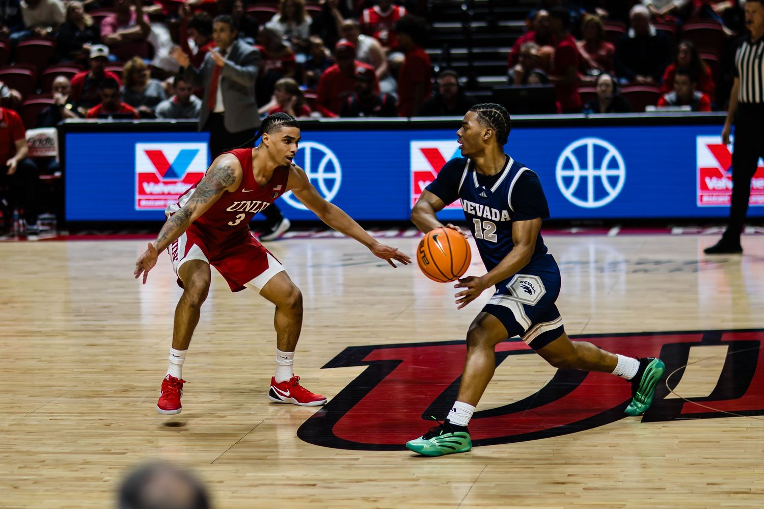 Two college basketball players compete on the court, one dribbling in a navy uniform while a red-uniformed defender guards closely.