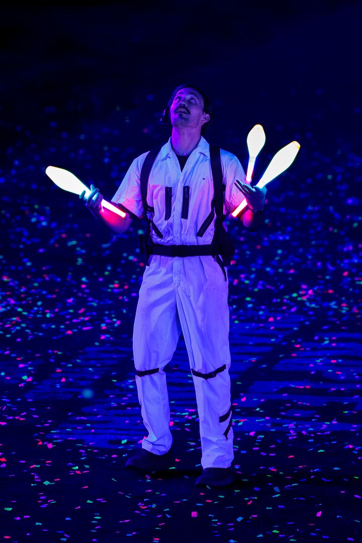 A juggler in a white jumpsuit performs with glowing LED clubs under neon blue stage lighting with colorful confetti.