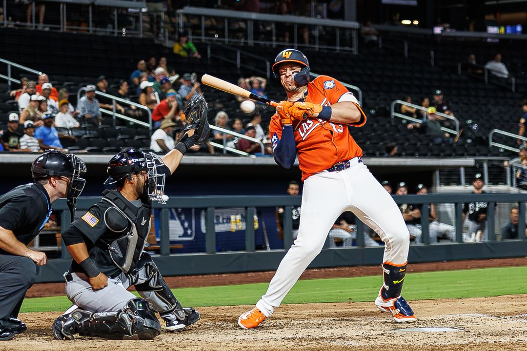 Baseball batter in orange uniform swings at pitch during night game with catcher and umpire behind home plate.