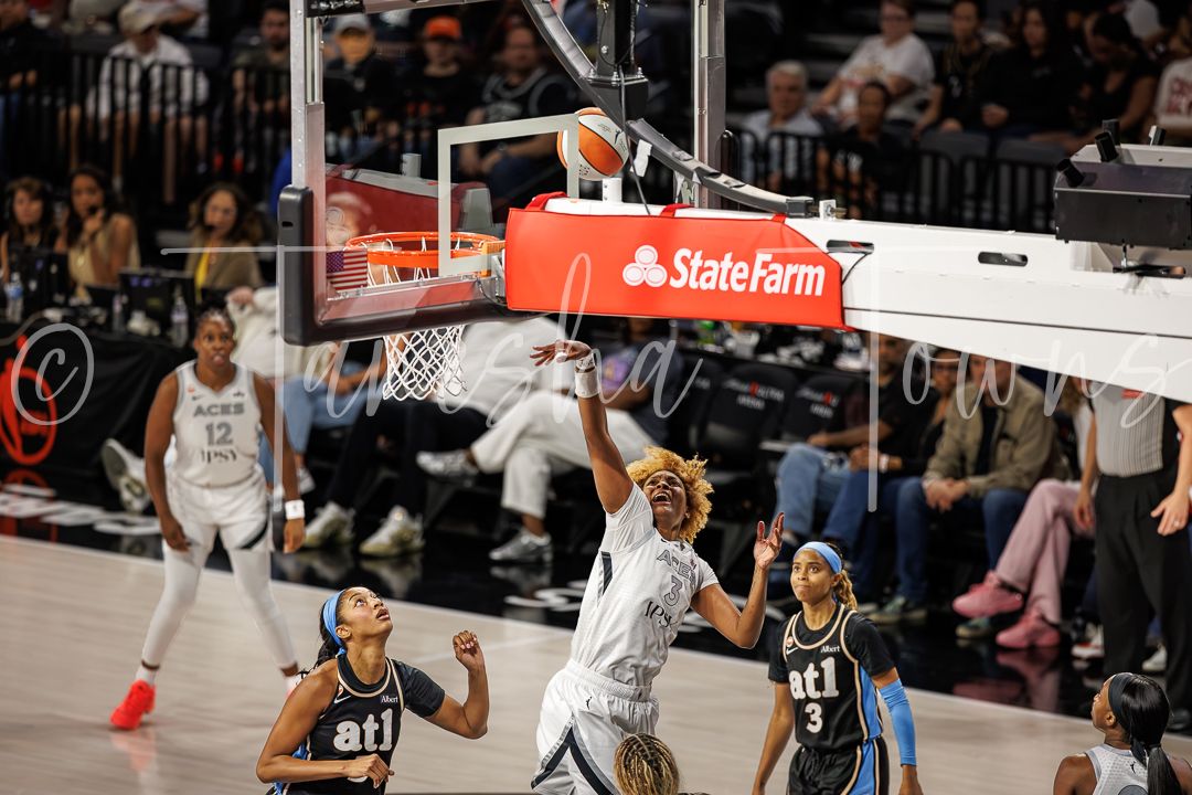 WNBA players compete near the basket during a game, with a State Farm sponsored backboard visible overhead.