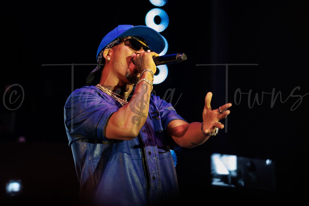 Male rapper performing on stage, wearing a blue cap and shirt, holding a microphone under dramatic concert lighting.