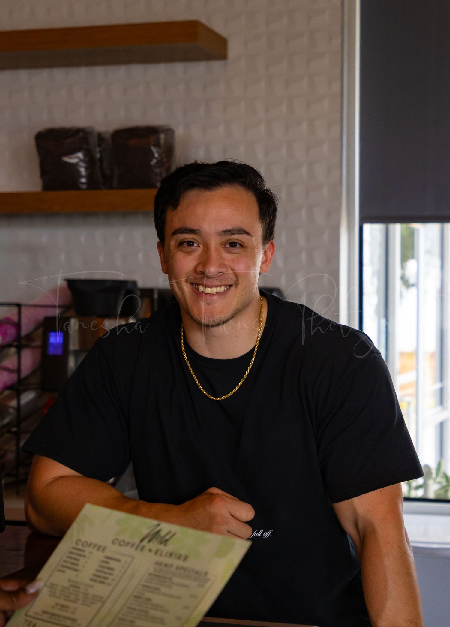 A person in a black t-shirt and gold chain smiles while looking at a menu in a casual restaurant setting.