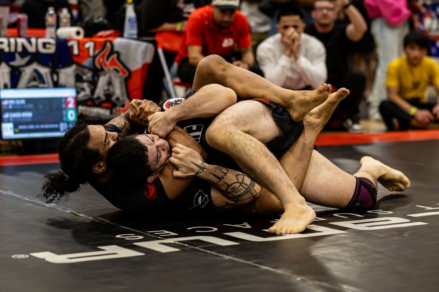 Two competitors grappling on the mat during a submission wrestling tournament, with spectators watching in the background.