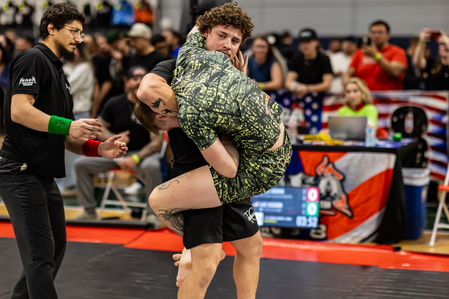 Two wrestlers compete in a grappling match at an indoor wrestling tournament with spectators watching.