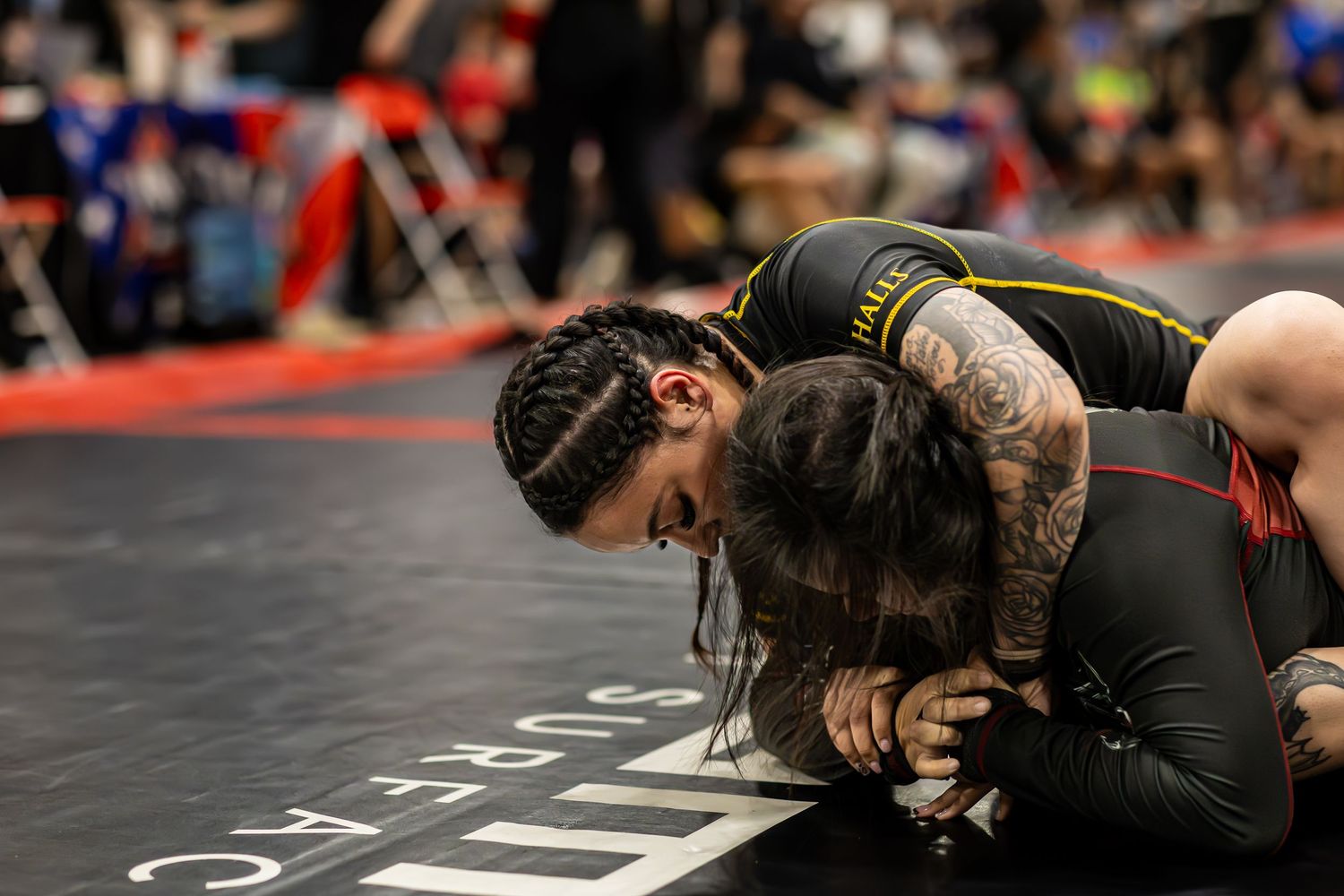 Two athletes competing in a Brazilian jiu-jitsu grappling match on a competition mat indoors.