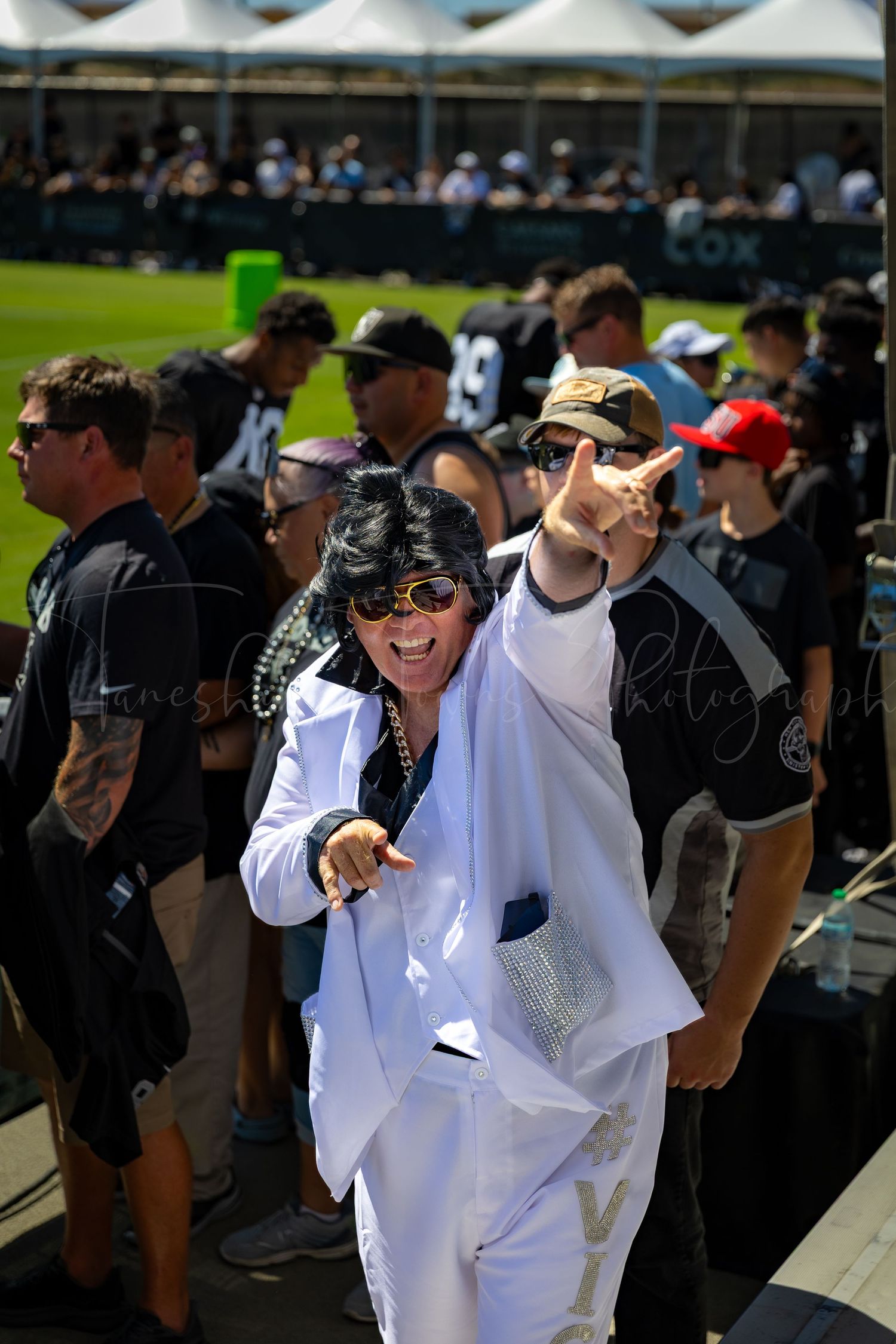 Fan in all-white outfit and sunglasses gestures enthusiastically at Raiders event.