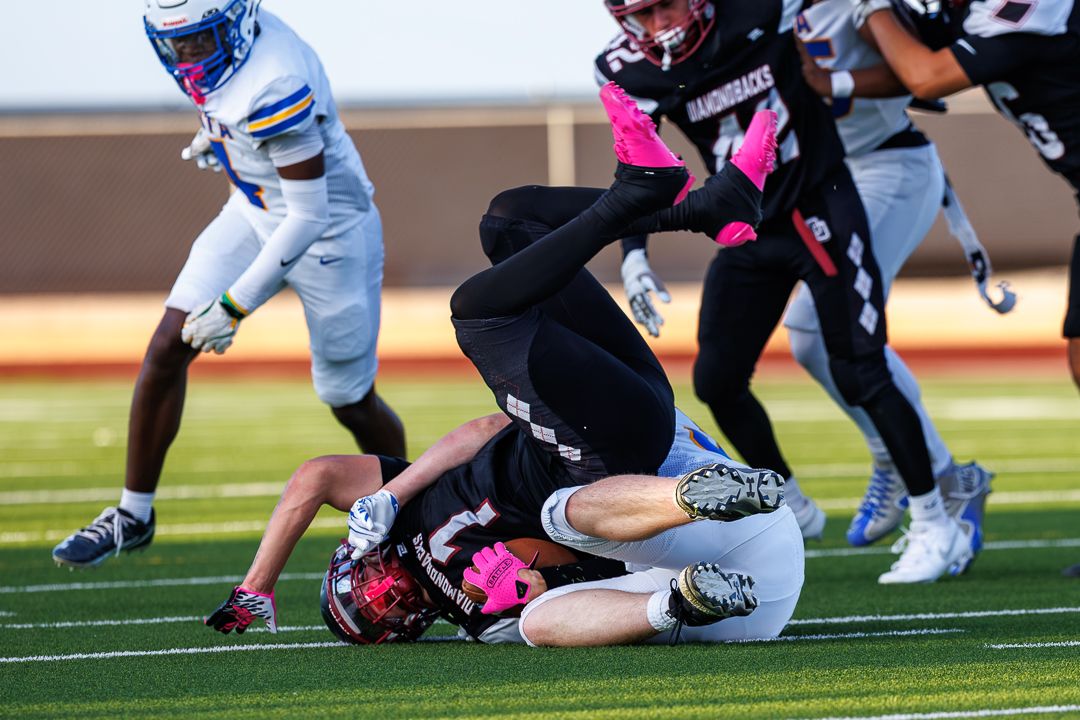 Football player tackled to the ground during a high school game with players in black and white uniforms on turf field.