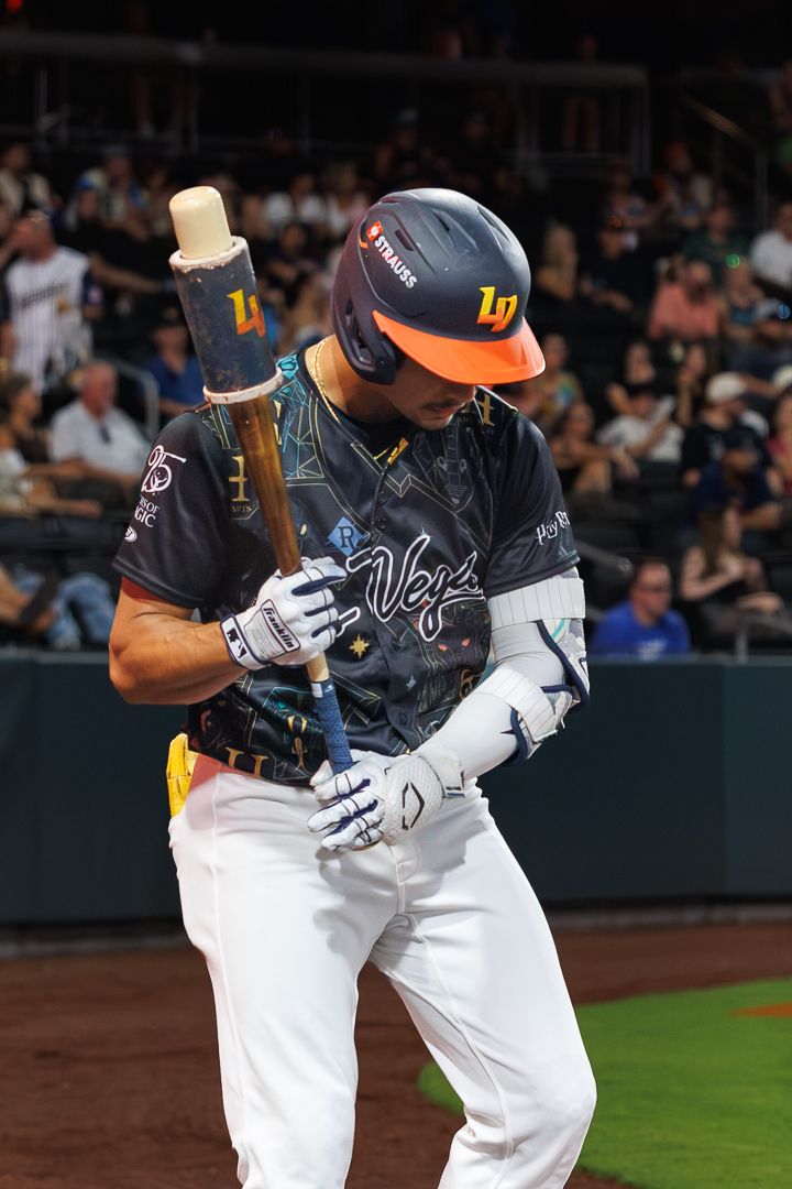 Las Vegas Aviators baseball player in dark uniform adjusts batting grip at home plate during night game.