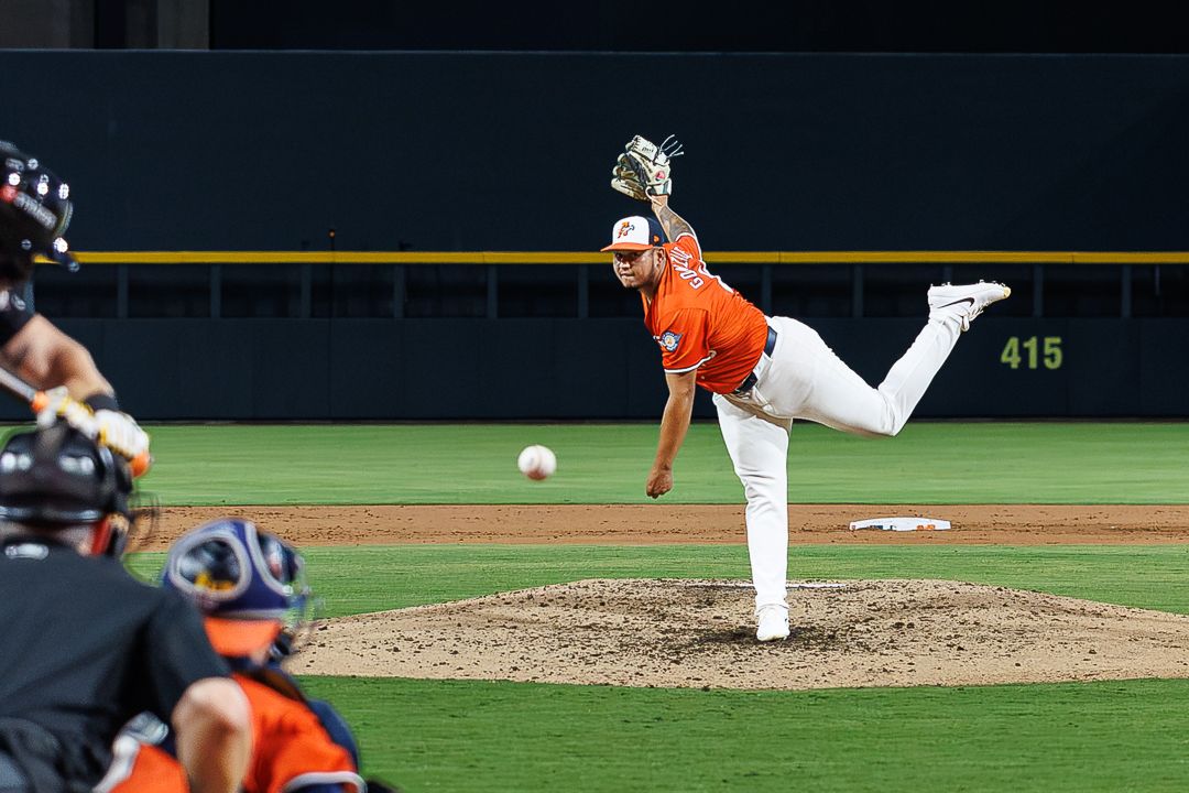 Baseball pitcher in orange uniform mid-throw on mound during night game with catcher and batter visible.