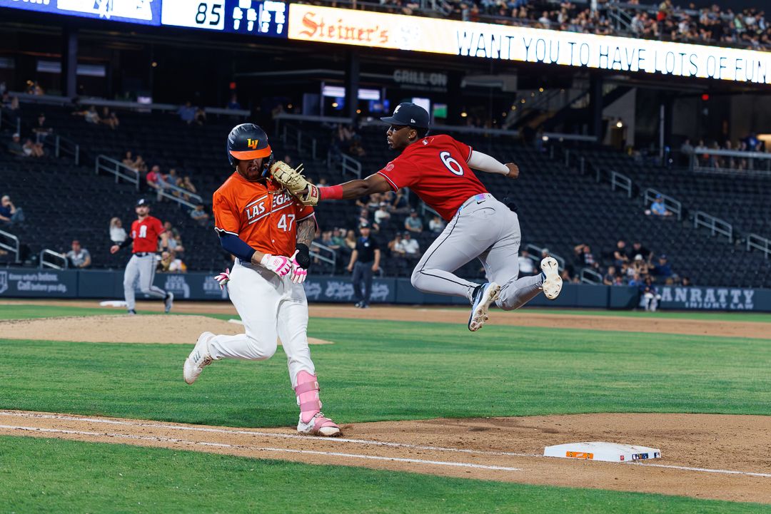 Baseball first baseman stretches to catch throw as runner arrives at first base during night game.