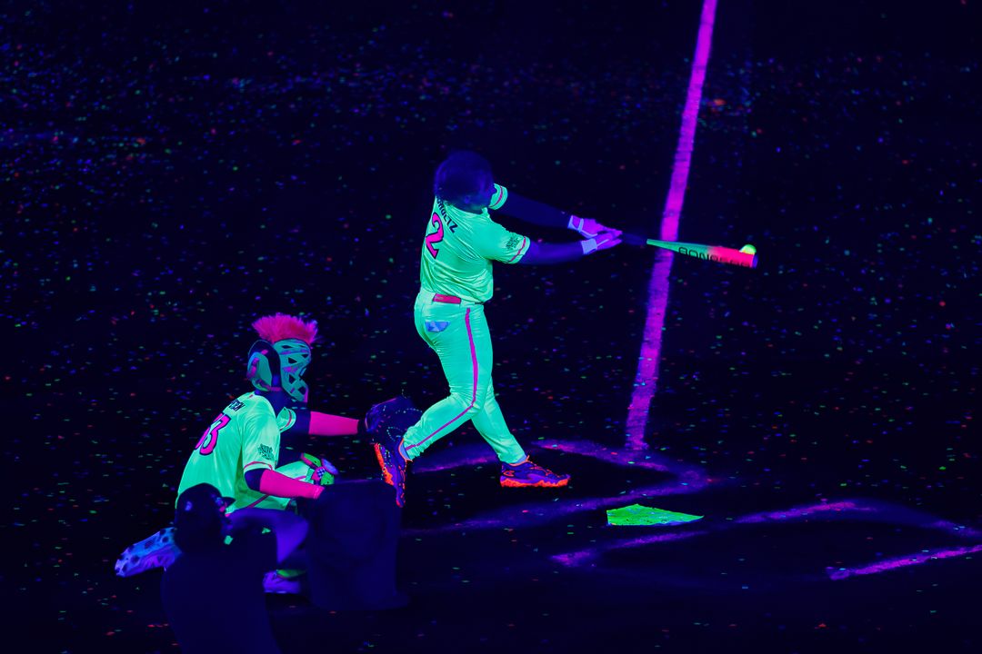 Neon glowing baseball batter swings at pitch while catcher crouches behind home plate under black light.
