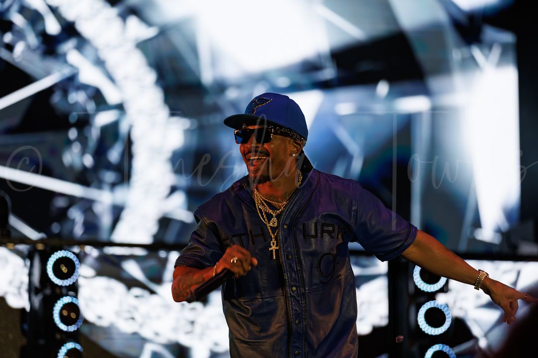 A performer in a blue shirt, cap, and sunglasses dances on a lit stage with glowing lights behind him.