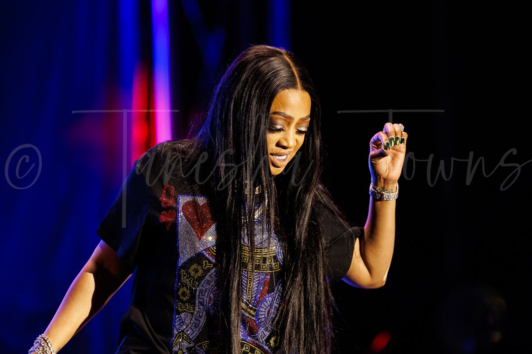 A female performer with long braids sings on stage under colorful lights, wearing a black graphic shirt.