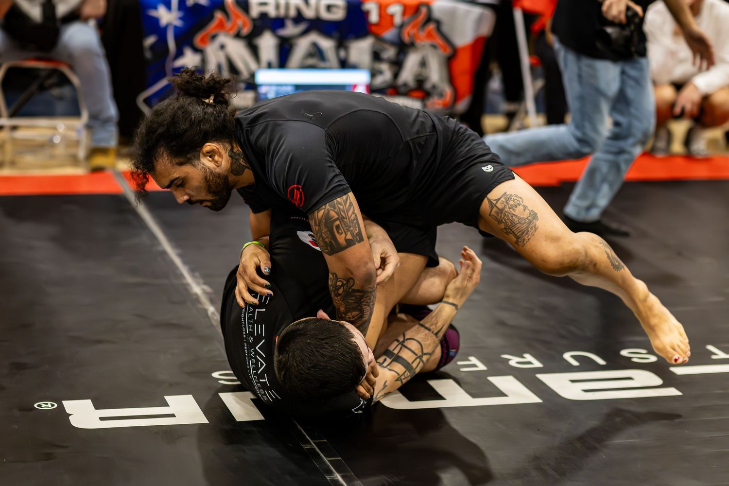 Two competitors grappling in a BJJ no-gi submission wrestling match at an indoor martial arts tournament.