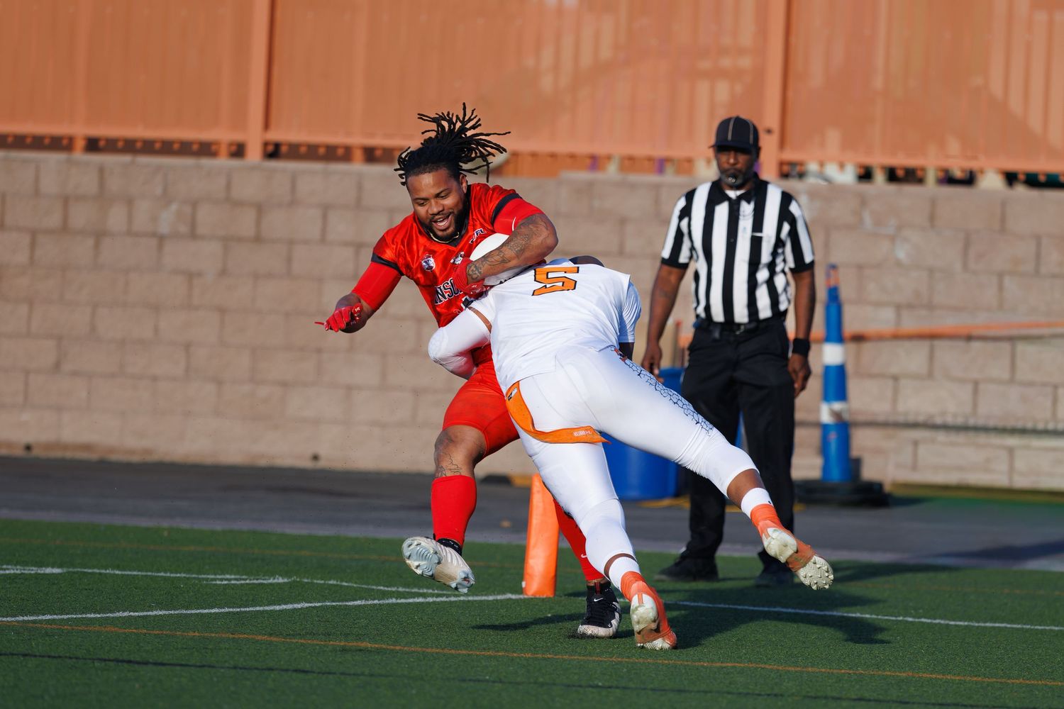 Two football players collide during a game, one in red and one in orange uniform, with a referee in background.