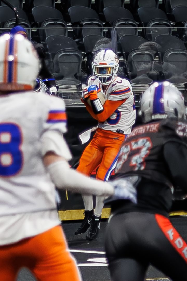 A football player in orange and blue uniform catches the ball during an indoor arena football game.