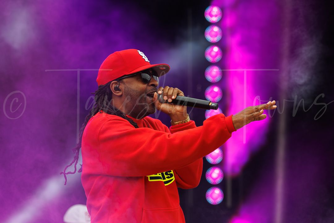 A performer in a red hoodie and cap sings into a microphone on a colorfully lit outdoor stage.