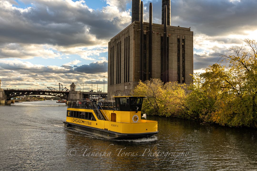 Dramatic cloudy skies over a waterfront industrial area with yellow barges and autumn trees along the riverbank.