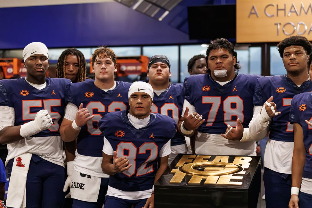 College football team in blue Jordan Brand uniforms poses together indoors near a championship trophy display.