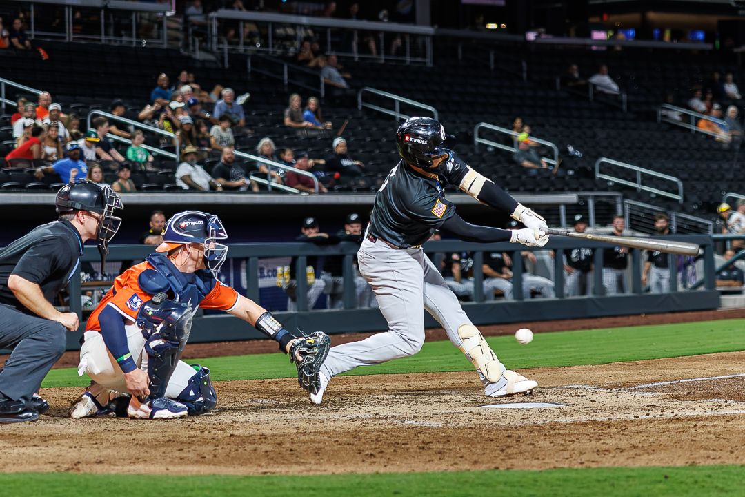 Baseball batter in dark uniform swings at pitch with catcher and umpire behind home plate.