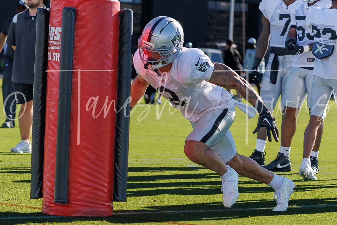 Football player in white uniform drives into a red blocking dummy during an outdoor practice drill on a sunny day.