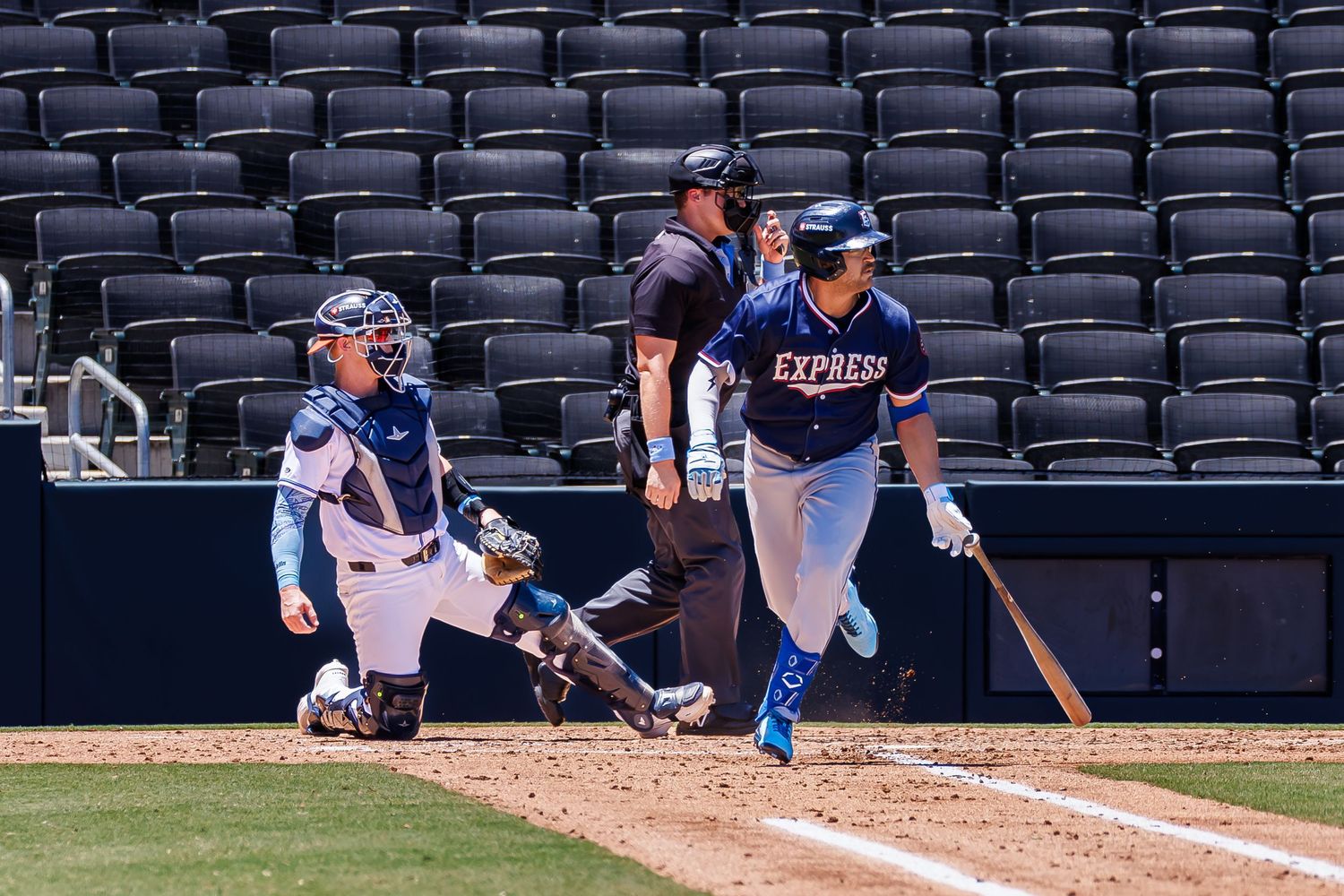Baseball batter in Express uniform swings at pitch with catcher and umpire behind home plate in empty stadium.