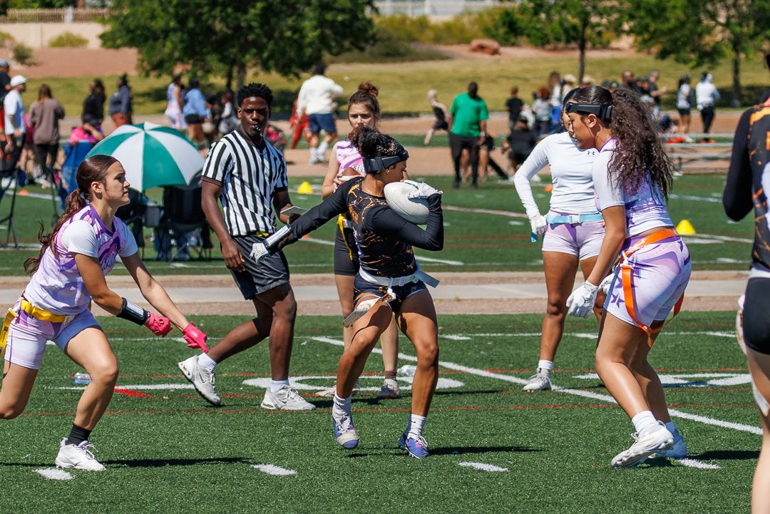 Players compete in a flag football game on a green turf field during a sunny outdoor sports event.
