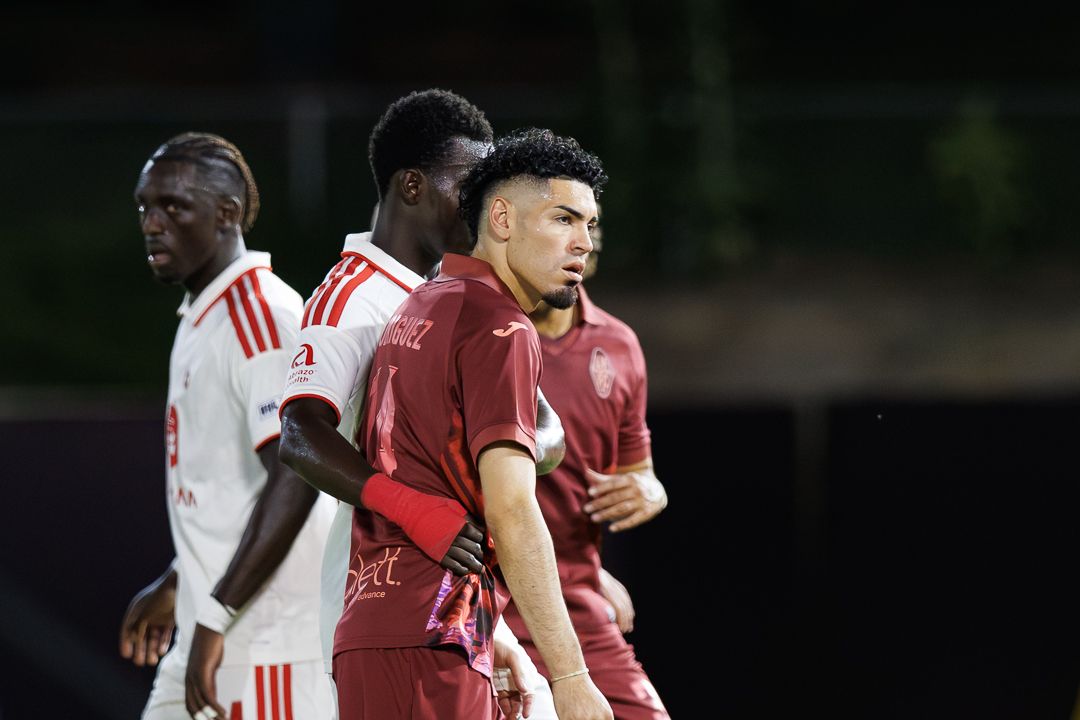 Two soccer players in maroon and white uniforms compete during a nighttime football match.