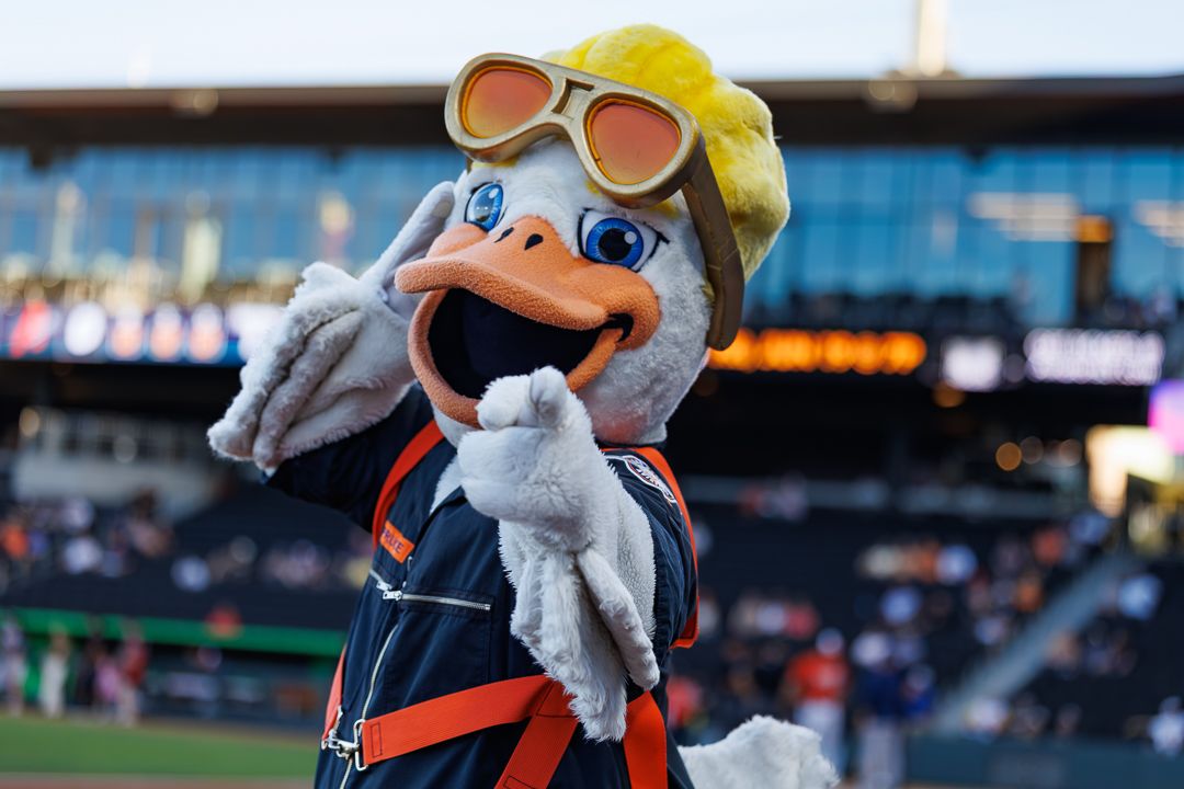 Baseball team mascot, a duck wearing goggles and overalls, poses energetically at the stadium.