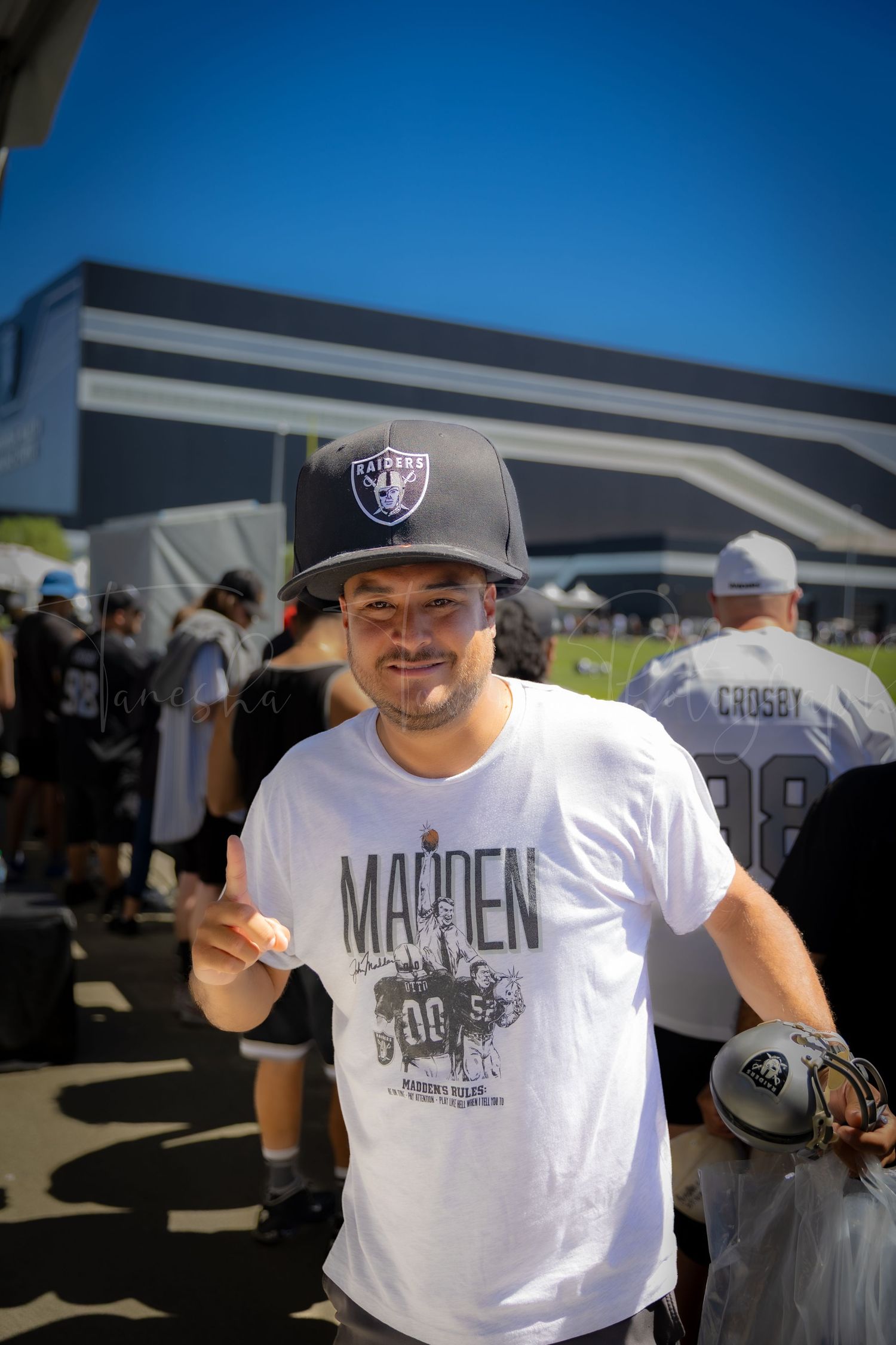 Raiders fan wearing a Madden t-shirt and team cap poses outside stadium during game day festivities.