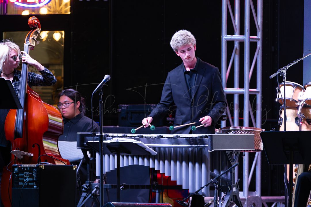 Jazz ensemble performers on stage with percussion instruments and lighting equipment visible.
