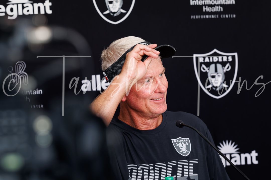 Raiders coach smiles at press conference in front of Allegiant and Raiders logos at Intermountain Health Performance Center.