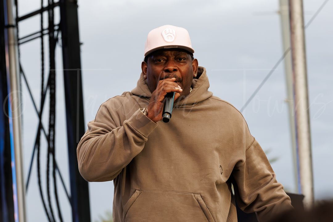 A man in a beige hoodie and white cap performs on stage holding a microphone at an outdoor concert.
